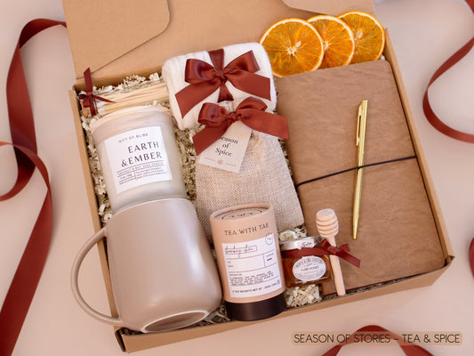 A warm and inviting gift box featuring a white ceramic mug, Earth & Ember candle, Bombay Chai tea and honey, a burlap sachet of fragrant seasonal spices for mulling or simmering, a vegan leather notebook and gold pen, soft socks, cinnamon sticks, and dried orange slices—designed for slow, cozy evenings.