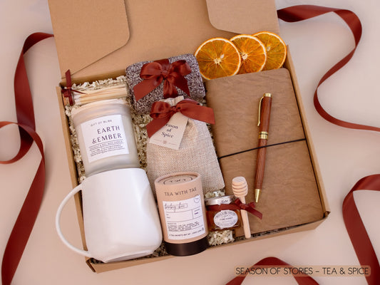 A warm and inviting gift box featuring a white ceramic mug, Earth & Ember candle, Bombay Chai tea and honey, a burlap sachet of fragrant seasonal spices for mulling or simmering, a vegan leather notebook and gold pen, soft socks, cinnamon sticks, and dried orange slices—designed for slow, cozy evenings.