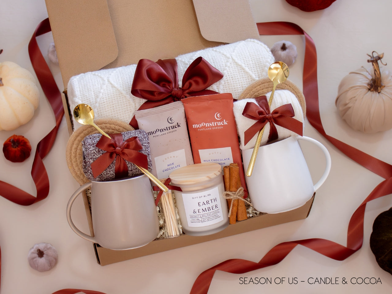 Gift box with 2 artisan hot cocoas, aromatherapy Earth & Ember candle, safety match jar, 2 minimalist mugs, 2 long golden spoons, 2 pairs of cozy socks, soft white blanket, 2 cotton yarn coasters, and cinnamon sticks, all decorated with satin ribbon in fall color, neatly arranged in a kraft gift box - a warm and comforting couples’ gift.