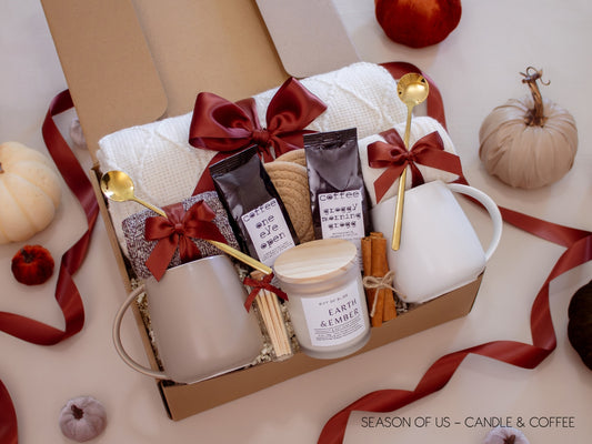 Gift box featuring 2 artisan coffee, aromatherapy Earth & Ember candle, safety match jar, 2 minimalist mugs, 2 long golden spoons, 2 pairs of cozy socks, soft white blanket, 2 cotton yarn coasters, and cinnamon sticks, all decorated with satin ribbon in fall color, neatly arranged in a kraft gift box - a cozy coffee and candlelight gift for couples.