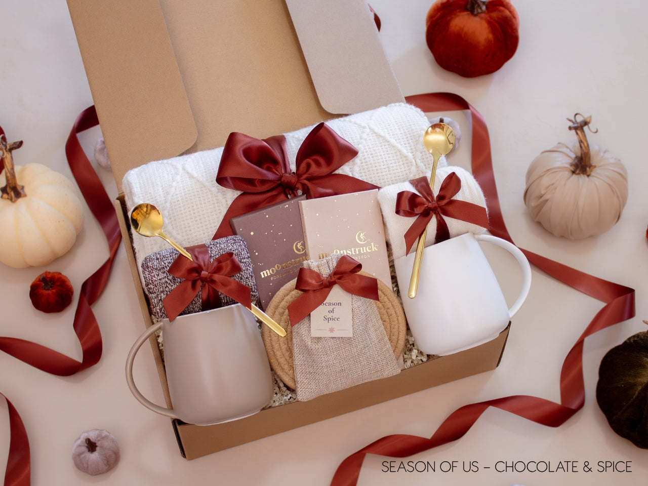 Couples’ gift box with decadent chocolate bars, aromatic spice bag for simmer pots or mulling, 2 minimalist mugs, 2 long golden spoons, 2 pairs of cozy socks, soft white blanket, and 2 cotton yarn coasters, all decorated with satin ribbon in fall color, neatly arranged in a kraft gift box — blending indulgence with cozy warmth.a