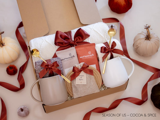 Gift box featuring rich cocoa, aromatic spice bag for simmer pots or mulling, 2 minimalist mugs, 2 long golden spoons, 2 pairs of cozy socks, soft white blanket, and 2 cotton yarn coasters, all decorated with satin ribbon in fall color, neatly arranged in a kraft gift box — a comforting couples’ gift for autumn nights.