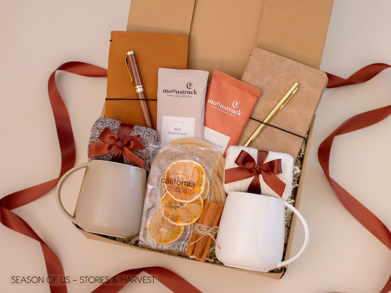 Couples’ gift box with journals, luxury wooden pen and gold pen, artisan cocoa, dried orange slices, cinnamon sticks, 2 minimalist mugs, 2 pairs of cozy socks, and 2 cotton yarn coasters, all decorated with satin ribbon in fall color, neatly arranged in a kraft gift box  — perfect for storytelling and fall evenings.