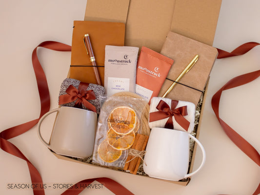 Couples’ gift box with journals, luxury wooden pen and gold pen, artisan cocoa, dried orange slices, cinnamon sticks, 2 minimalist mugs, 2 pairs of cozy socks, and 2 cotton yarn coasters, all decorated with satin ribbon in fall color, neatly arranged in a kraft gift box  — perfect for storytelling and fall evenings.