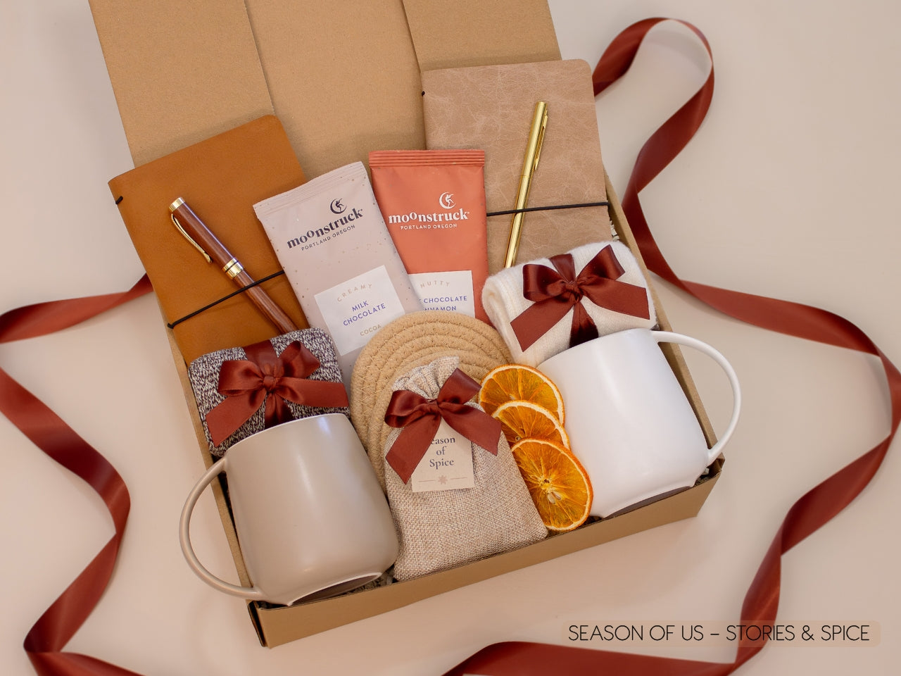 Gift box with journals, luxury wooden pen and gold pen, artisan cocoa, aromatic spice bag for simmer pots or mulling, 2 minimalist mugs, 2 pairs of cozy socks, and 2 cotton yarn coasters, all decorated with satin ribbon in fall color, neatly arranged in a kraft gift box — a thoughtful couples’ gift for reflection and connection.
