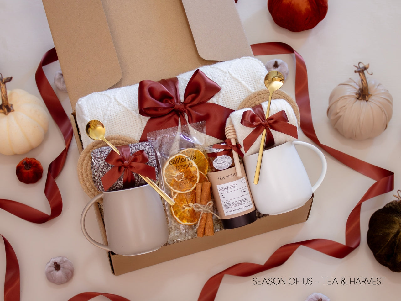 Gift box with fragrant tea, pure honey, wooden dipper, 2 minimalist mugs, 2 long golden spoons, 2 pairs of cozy socks, soft white blanket, 2 cotton yarn coasters, dried orange slices, and cinnamon sticks, all decorated with satin ribbon in fall color, neatly arranged in a kraft gift box — a simple and cozy tea-themed couples’ gift for relaxation and connection.