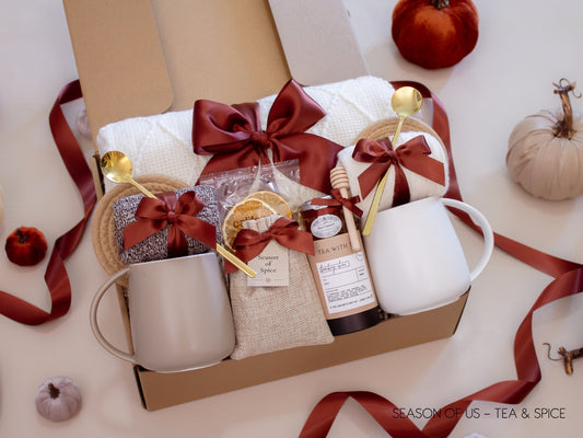 Gift box with artisan tea, pure honey, wooden dipper, dried orange slises, aromatic spice bag for simmer pots or mulling, 2 minimalist mugs, 2 long golden spoons, 2 pairs of cozy socks, soft white blanket, and 2 cotton yarn coasters, all decorated with satin ribbon in fall color, neatly arranged in a kraft gift box — a fall-inspired couples’ gift.