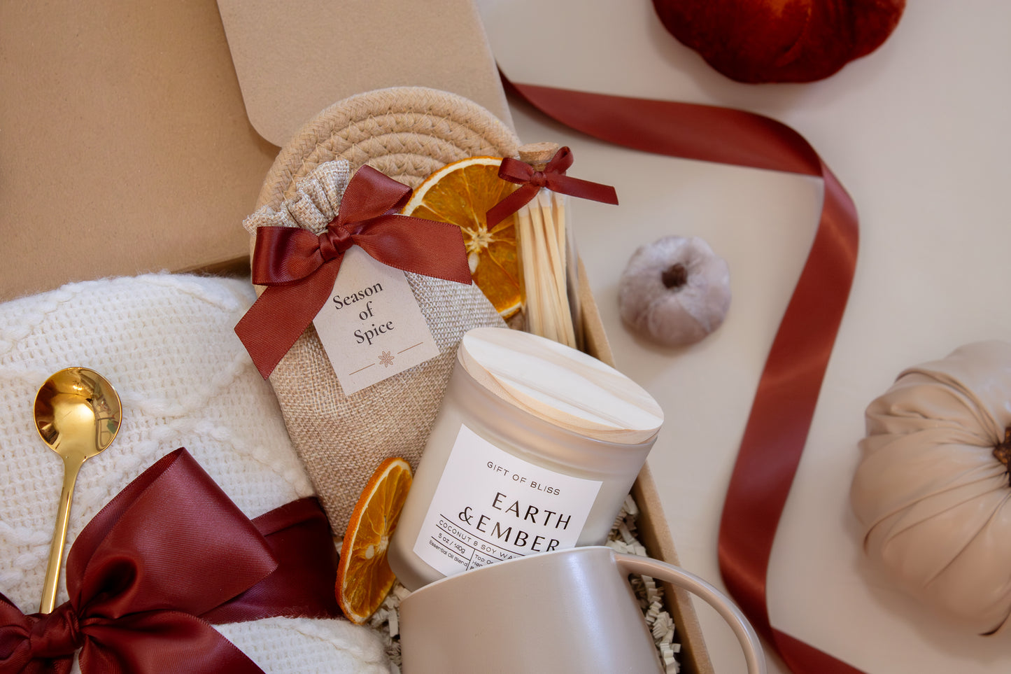 A warm gift box with a plush white knit blanket tied in a burgundy satin bow, a Season of Spice sachet with seasonal spices for mulling or simmering, dried oranges, a golden spoon, Earth & Ember candle with matches, and a taupe ceramic mug—perfect for a cozy autumn night.