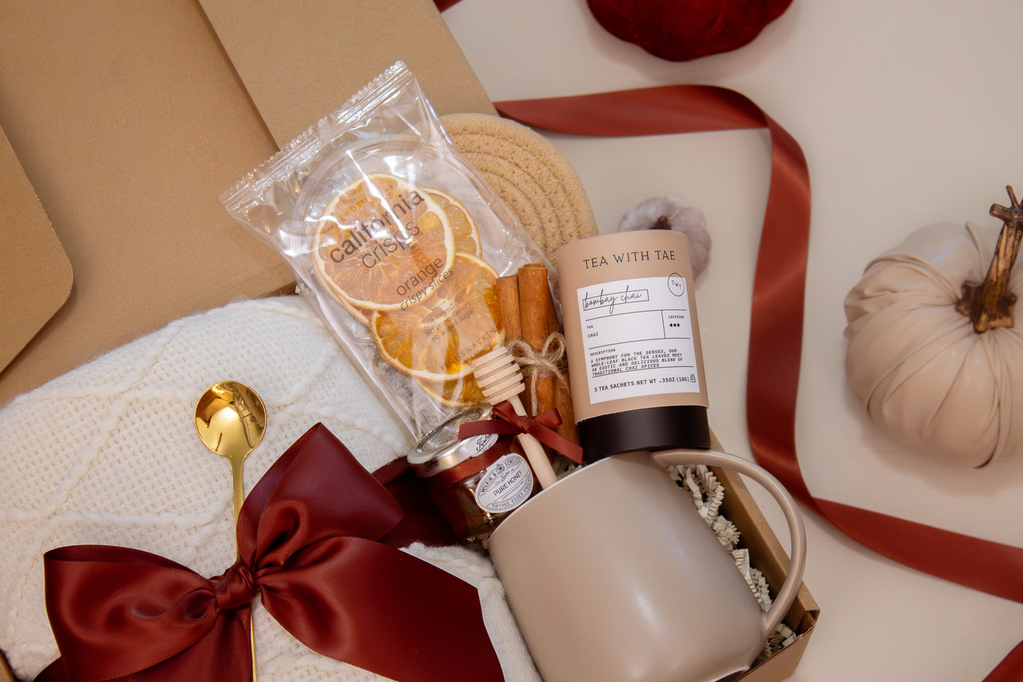 A harvest-inspired gift box with a soft white knit blanket tied with a burgundy satin bow, Bombay Chai tea, honey jar with dipper, orange crisps, Earth & Ember candle with matches, and a taupe ceramic mug—bringing the flavors and comfort of fall together.