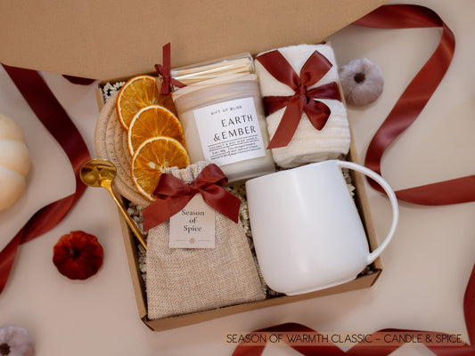 A rustic gift box with a taupe mug, Earth & Ember candle, a burlap sachet of fragrant seasonal spices for mulling or simmering, soft socks, a golden spoon, cinnamon sticks, and dried orange slices—bringing a touch of autumn spice and cozy warmth.
