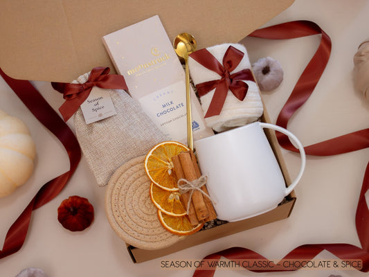 A festive gift box with a taupe mug, creamy Moonstruck milk chocolate, a burlap sachet of seasonal spices for mulling or simmering, soft socks, golden spoon, cinnamon sticks, and dried orange slices—ideal for savoring sweet, spiced evenings.