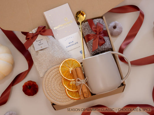 A festive gift box with a taupe mug, creamy Moonstruck milk chocolate, a burlap sachet of seasonal spices for mulling or simmering, soft socks, golden spoon, cinnamon sticks, and dried orange slices—ideal for savoring sweet, spiced evenings.