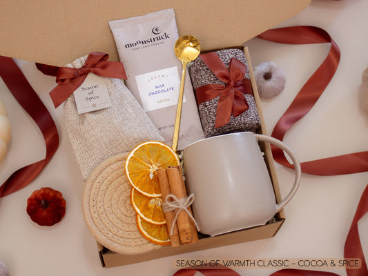 A cozy kraft box with a taupe mug, Moonstruck cocoa bar, burlap sachet of seasonal spices for mulling or simmering, soft socks, golden spoon, cinnamon sticks, and dried orange slices—created to wrap chilly nights in comforting chocolate and spice.