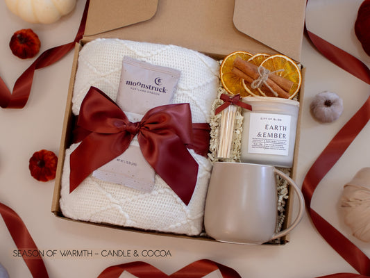 A cozy gift box with a soft white knit blanket tied with a burgundy satin bow, creamy milk chocolate cocoa, a warm Earth & Ember candle with matches, dried orange slices, and a taupe ceramic mug—everything for a comforting cocoa night.