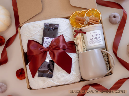 A beautifully packed gift box with a soft white knit blanket tied in a burgundy bow, rich groggy morning coffee, Earth & Ember candle with matches, dried orange slices, and a taupe ceramic mug—perfect for a cozy coffee moment.