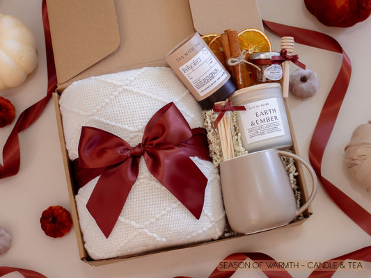 A warm and elegant gift box featuring a plush white knit blanket wrapped with a burgundy bow, artisan chai tea with honey and dipper, Earth & Ember candle with matches, dried orange slices, and a taupe ceramic mug for a soothing tea break.
