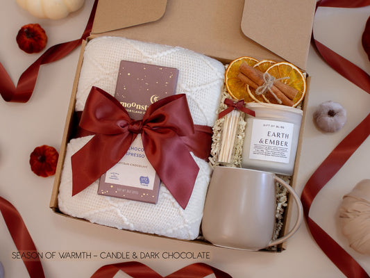 An inviting gift box with a white knit blanket tied with a burgundy satin bow, artisan chocolate, Earth & Ember candle with matches, dried orange slices, and a taupe ceramic mug—crafted for a sweet and cozy evening.