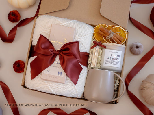 An inviting gift box with a white knit blanket tied with a burgundy satin bow, artisan chocolate, Earth & Ember candle with matches, dried orange slices, and a taupe ceramic mug—crafted for a sweet and cozy evening.