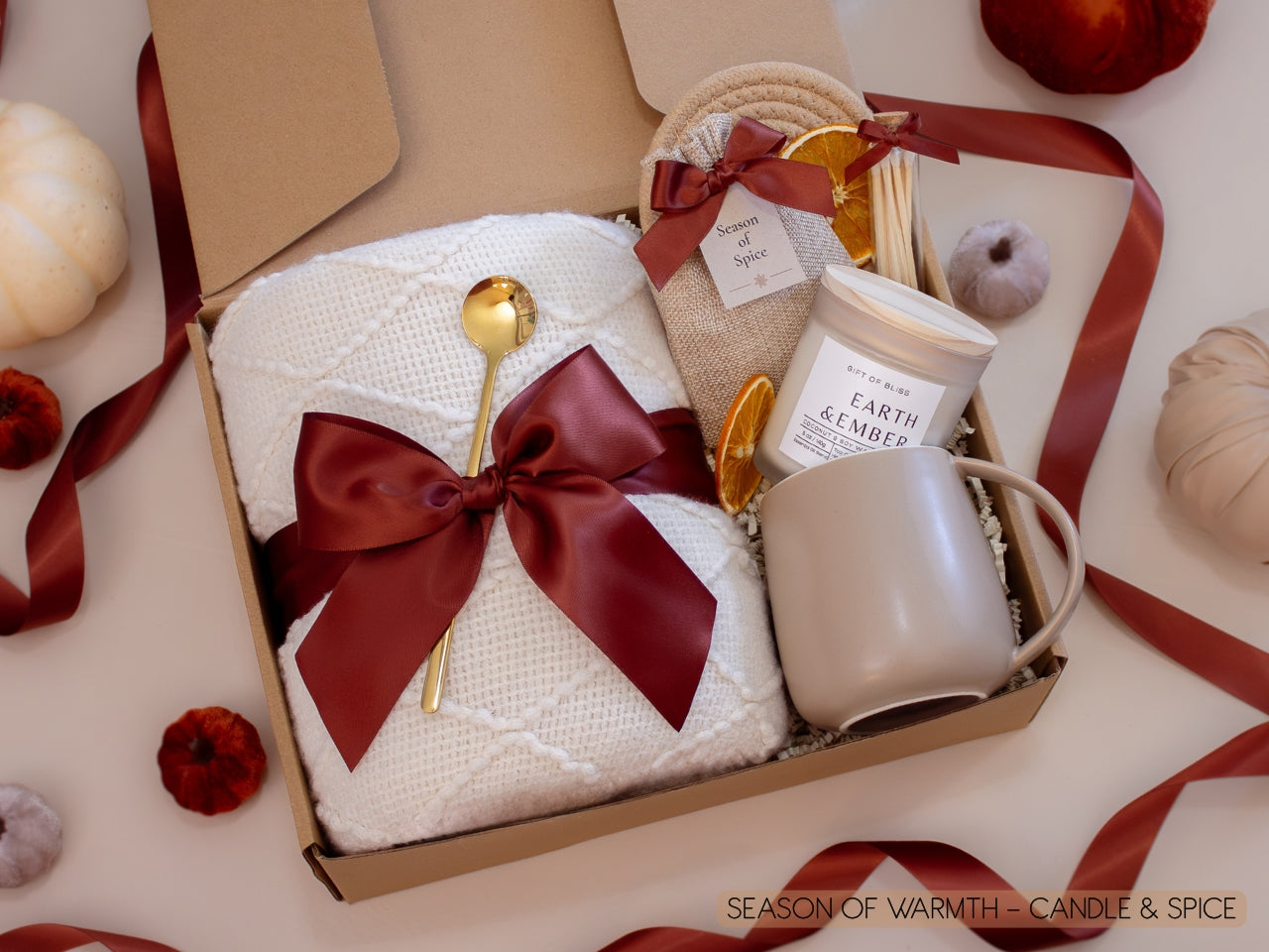 A warm gift box with a plush white knit blanket tied in a burgundy satin bow, a Season of Spice sachet with seasonal spices for mulling or simmering, dried oranges, a golden spoon, Earth & Ember candle with matches, and a taupe ceramic mug—perfect for a cozy autumn night.