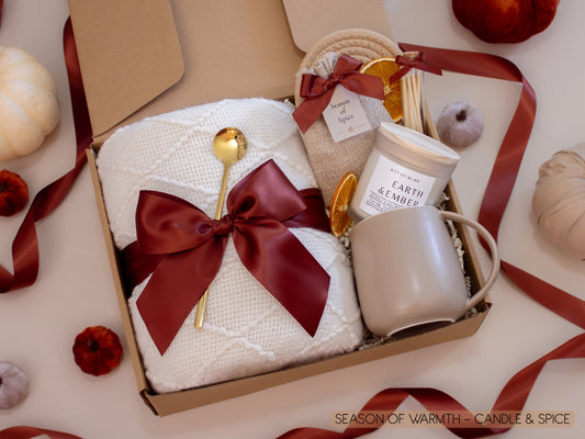 A warm gift box with a plush white knit blanket tied in a burgundy satin bow, a Season of Spice sachet with seasonal spices for mulling or simmering, dried oranges, a golden spoon, Earth & Ember candle with matches, and a taupe ceramic mug—perfect for a cozy autumn night.