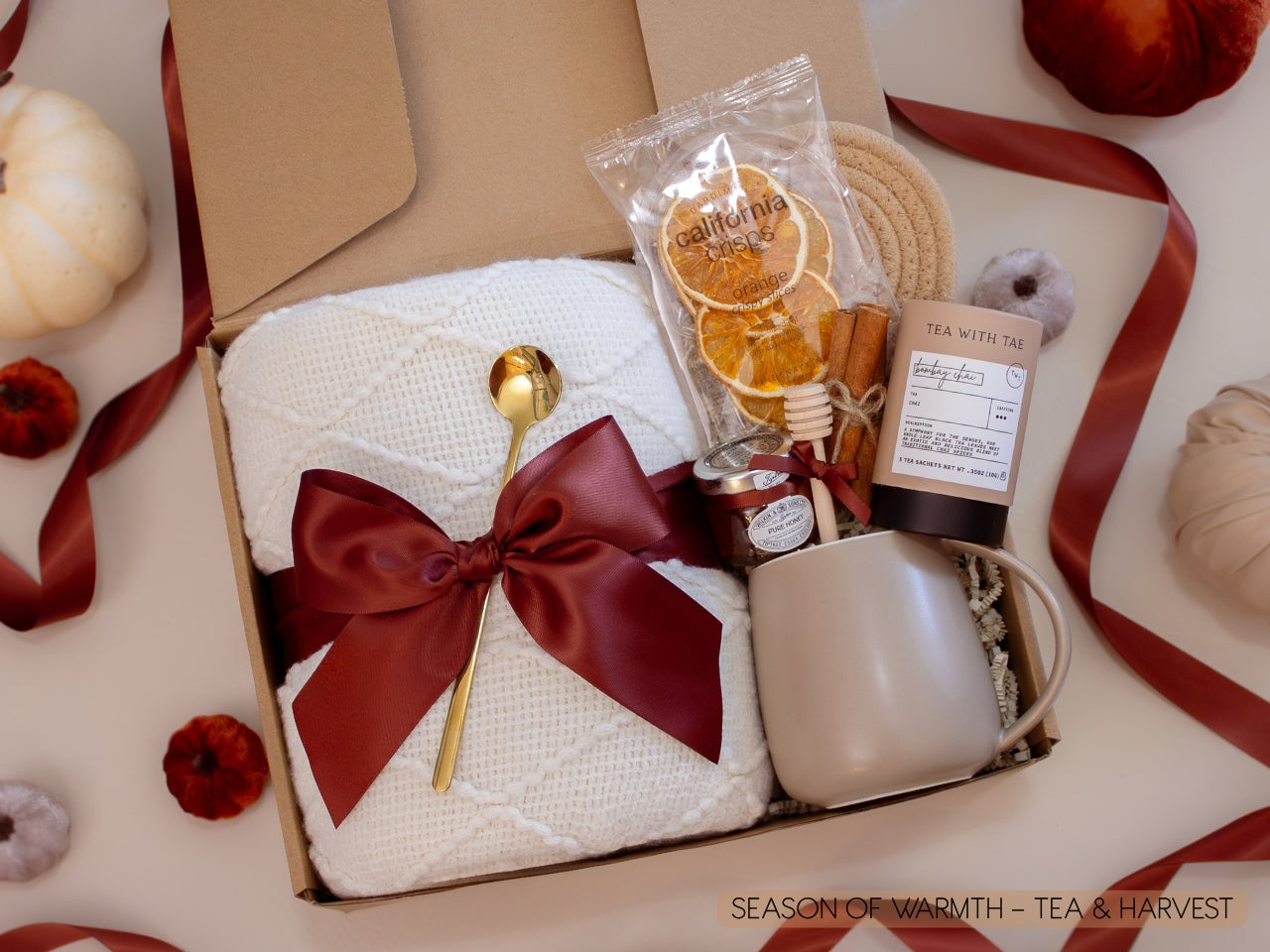 A harvest-inspired gift box with a soft white knit blanket tied with a burgundy satin bow, Bombay Chai tea, honey jar with dipper, orange crisps, Earth & Ember candle with matches, and a taupe ceramic mug—bringing the flavors and comfort of fall together.