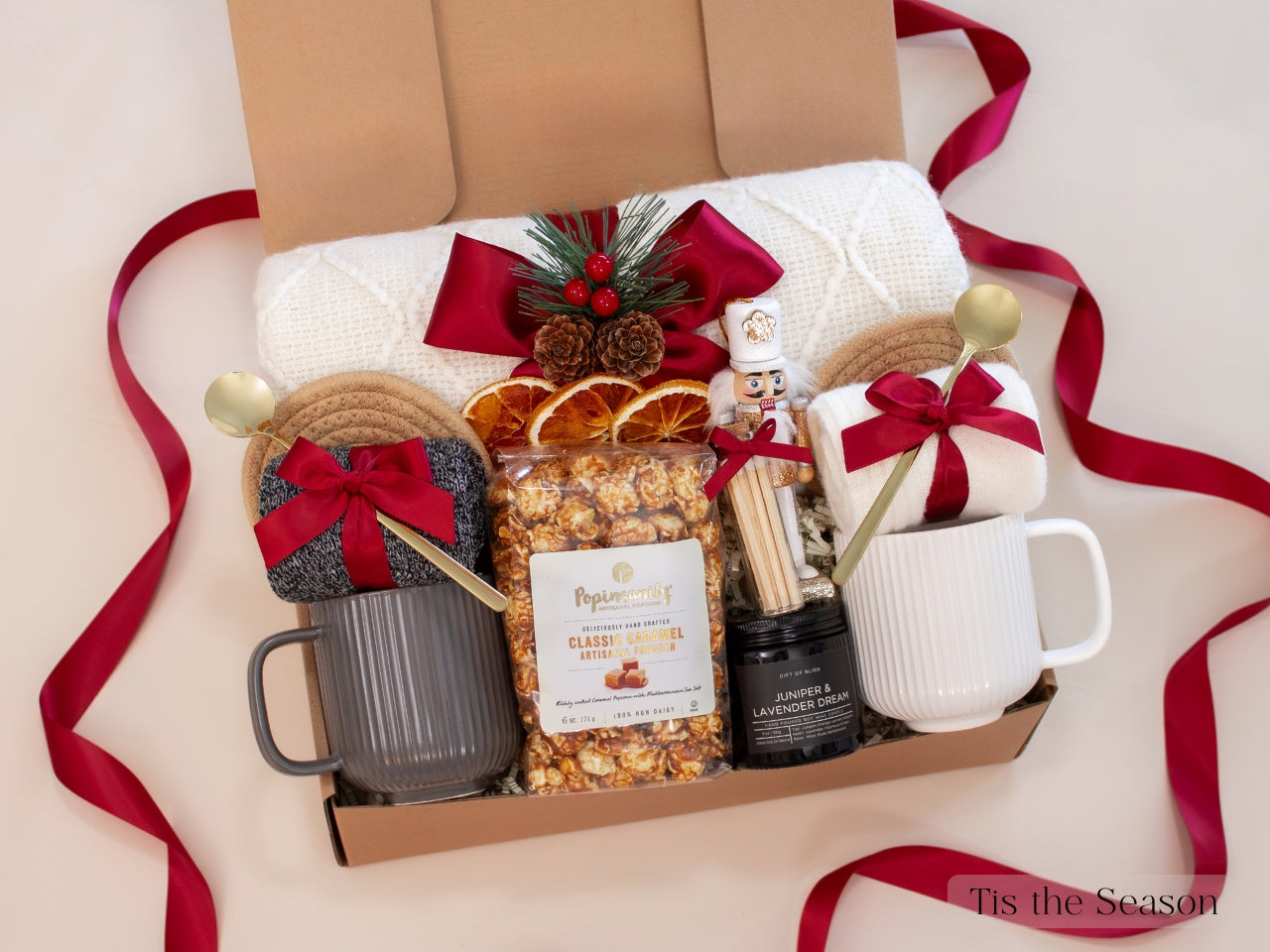 Cozy and festive Christmas gift box with gray and white mugs, caramel popcorn, festive pine and berry decor, nutcracker, red ribbons, and knit blanket — a warm and comforting holiday gift for couples.