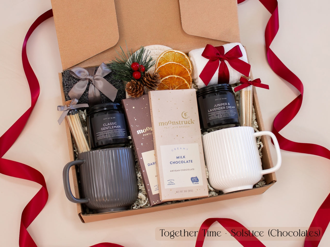 Luxurious holiday gift box for couples featuring gray and white ribbed mugs, artisan chocolate bars, pinecone and dried orange decor, cozy socks, and festive red ribbons — a warm and modern Christmas gift set.