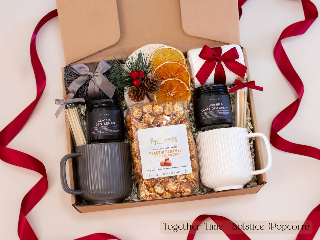 Luxurious holiday gift box for couples featuring gray and white ribbed mugs, caramel popcorn, pinecone and dried orange decor, cozy socks, and festive red ribbons — a warm and modern Christmas gift set.