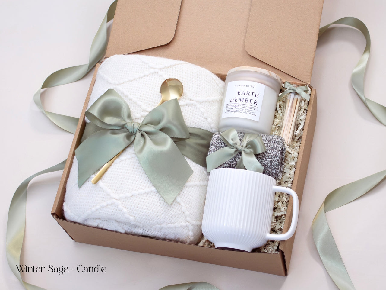 Minimalist winter gift box with a white mug, ribbed candle, cozy socks tied in sage ribbon, and a plush white blanket accented by a satin green bow.