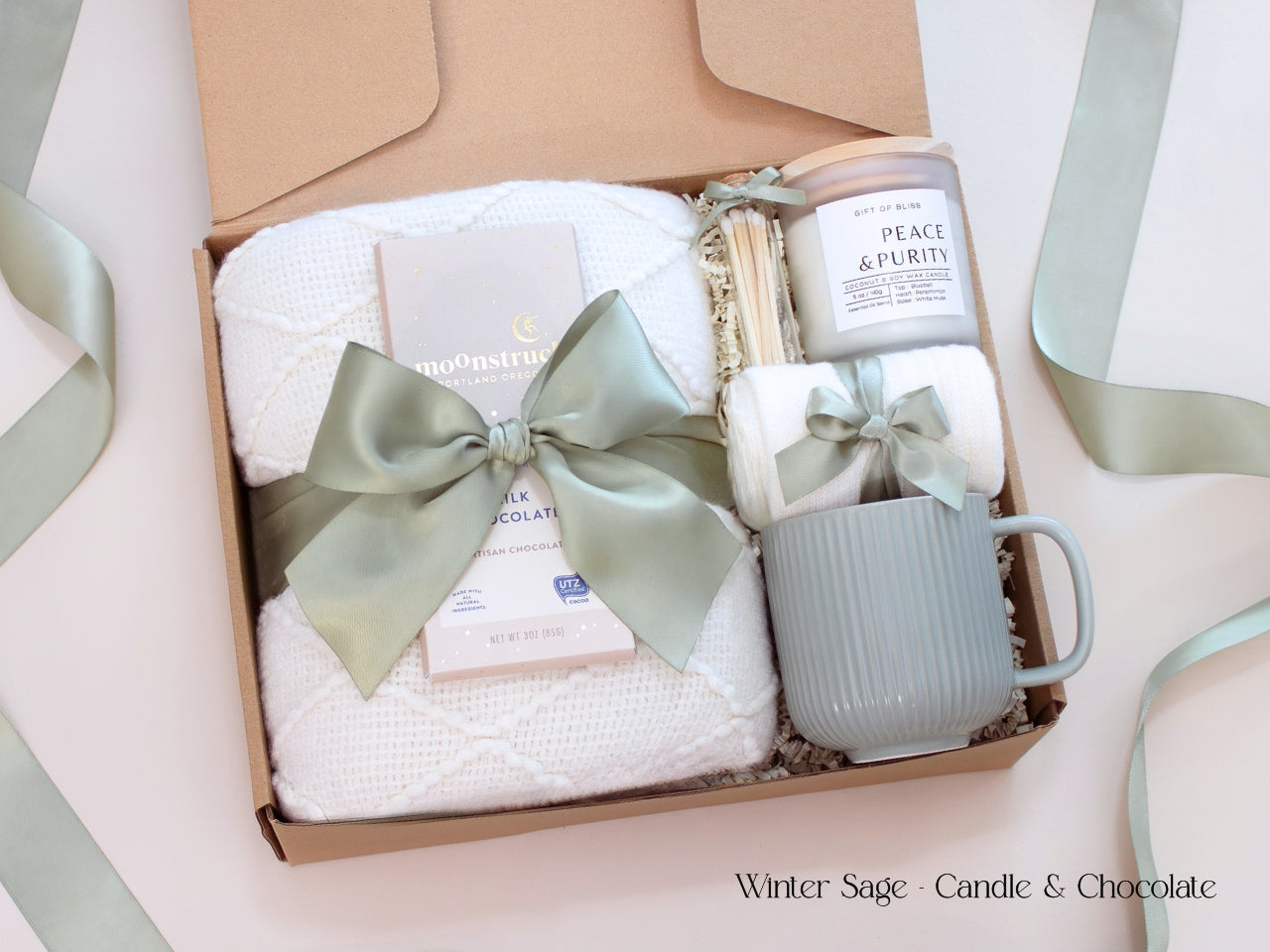 A serene gift box featuring a white textured blanket, a ribbed white mug with cozy socks tied in sage ribbon, a coconut soy wax candle, and a Moonstruck chocolate bar, all presented with a soft sage bow and minimalist winter aesthetic.