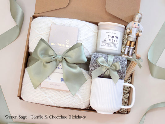 A holiday-inspired gift box with a cozy white blanket, white mug, socks tied in sage ribbon, Moonstruck chocolate, and a candle labeled “Earth & Ember,” accented with a golden nutcracker and winter greenery.