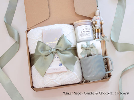 A holiday-inspired gift box with a cozy white blanket, white mug, socks tied in sage ribbon, Moonstruck chocolate, and a candle labeled “Earth & Ember,” accented with a golden nutcracker and winter greenery.