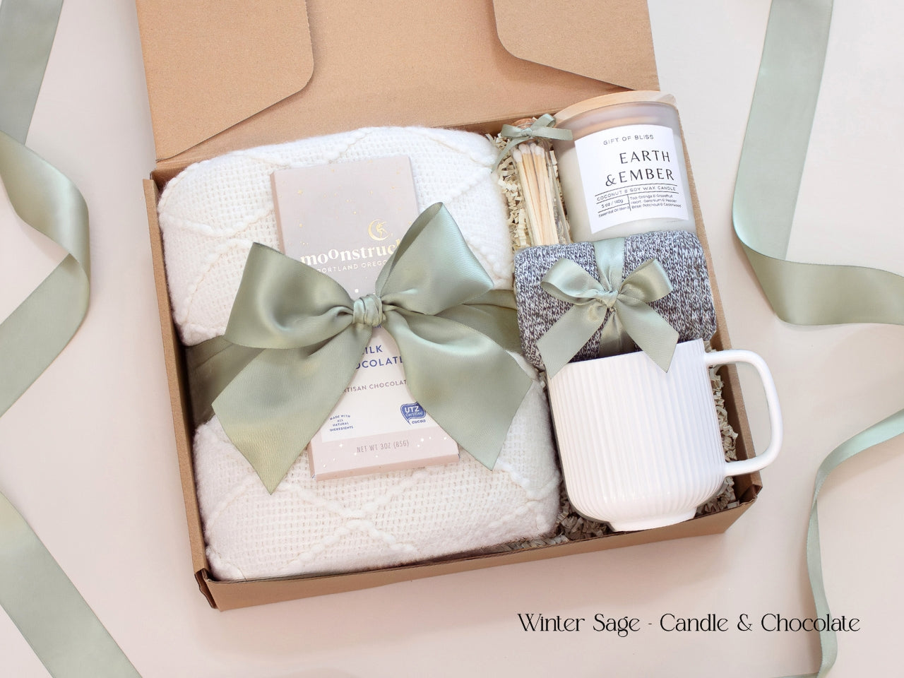 A serene gift box featuring a white textured blanket, a ribbed white mug with cozy socks tied in sage ribbon, a coconut soy wax candle, and a Moonstruck chocolate bar, all presented with a soft sage bow and minimalist winter aesthetic.