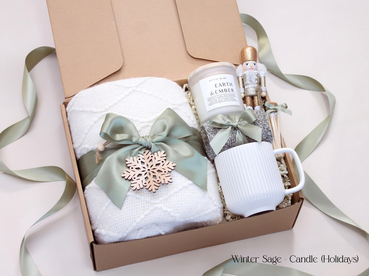 Elegant winter holiday gift box featuring a white blanket tied with a sage bow and wooden snowflake, a white mug, candle, socks, and a gold-accented nutcracker figurine.