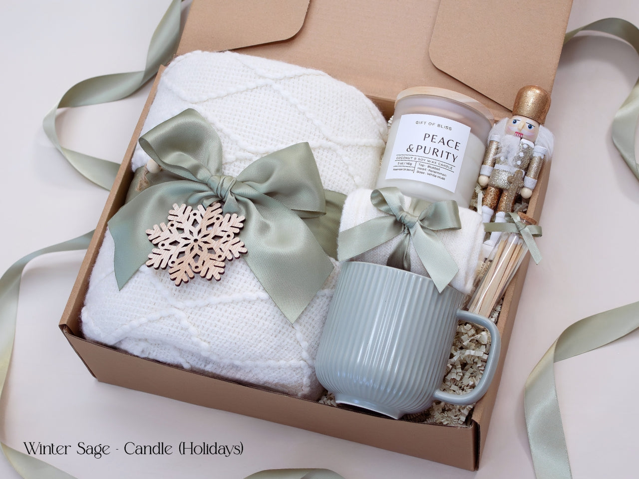 Elegant winter holiday gift box featuring a white blanket tied with a sage bow and wooden snowflake, a white mug, candle, socks, and a gold-accented nutcracker figurine.
