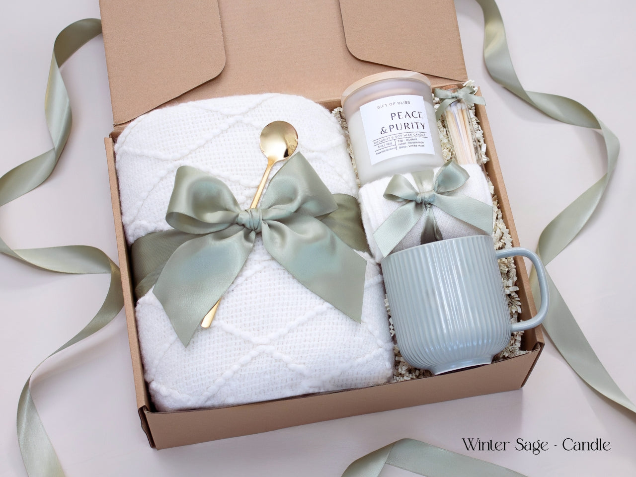 Minimalist winter gift box with a white mug, ribbed candle, cozy socks tied in sage ribbon, and a plush white blanket accented by a satin green bow.