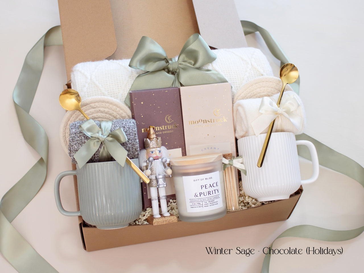 A beautifully arranged holiday gift box for couples from the Winter Sage Collection featuring a white knit blanket, two ribbed mugs in sage green and white, cozy socks tied with satin bows, Moonstruck artisan chocolate bars, a Peace & Purity candle, and a small nutcracker accent. The box is tied with a soft sage ribbon, creating a calm and elegant holiday feel.
