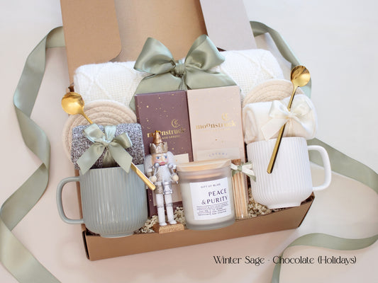 A beautifully arranged holiday gift box for couples from the Winter Sage Collection featuring a white knit blanket, two ribbed mugs in sage green and white, cozy socks tied with satin bows, Moonstruck artisan chocolate bars, a Peace & Purity candle, and a small nutcracker accent. The box is tied with a soft sage ribbon, creating a calm and elegant holiday feel.