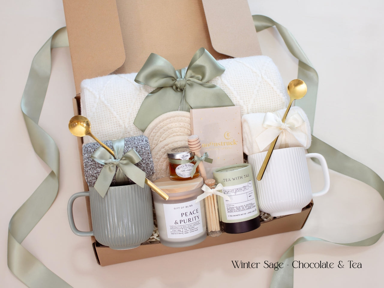A cozy Winter Sage gift box for couples with a white knit blanket, two mugs, soft socks, Moonstruck chocolate, Tea with Tae tea tin, pure honey, and a Peace & Purity candle. Designed with a calming green and cream palette and finished with a satin sage ribbon — perfect for thoughtful gifting year-round.