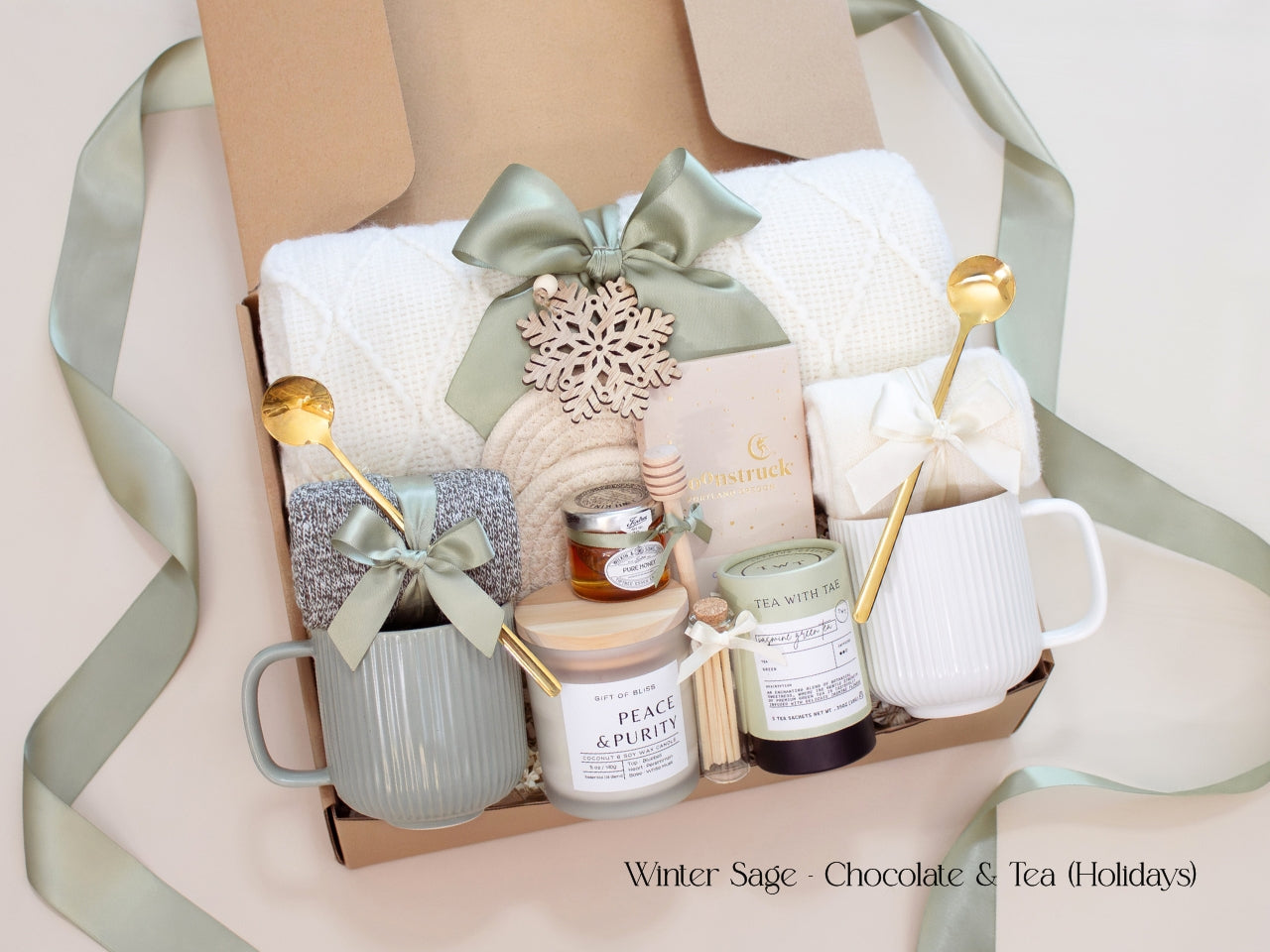An elegant Winter Sage holiday gift box for couples including a white knit blanket, two mugs, socks tied with satin bows, Moonstruck chocolate, Tea with Tae artisan tea tin, pure honey jar, a Peace & Purity candle, and a wooden snowflake ornament. Styled with sage ribbon for a warm, festive, and cozy presentation.