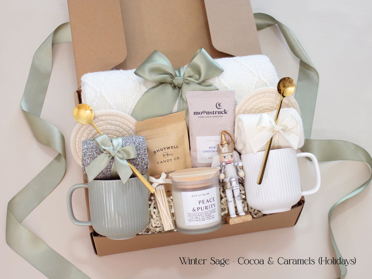 A luxurious holiday gift box for couples featuring Moonstruck chocolate, Shotwell caramels, a Peace & Purity candle, cozy socks, mugs in sage green and white, a white knit blanket, and a small nutcracker accent. Wrapped with a sage green ribbon, exuding warmth and festive charm.