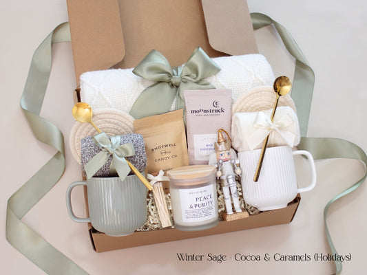 A luxurious holiday gift box for couples featuring Moonstruck chocolate, Shotwell caramels, a Peace & Purity candle, cozy socks, mugs in sage green and white, a white knit blanket, and a small nutcracker accent. Wrapped with a sage green ribbon, exuding warmth and festive charm.