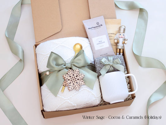 A holiday gift box for her featuring a white blanket tied with sage ribbon and wooden snowflake, a white mug, socks, hot cocoa, and small caramel packet with a nutcracker accent.