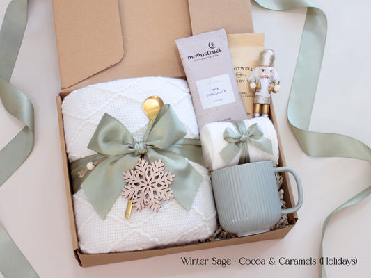 A holiday gift box for her featuring a white blanket tied with sage ribbon and wooden snowflake, a white mug, socks, hot cocoa, and small caramel packet with a nutcracker accent.