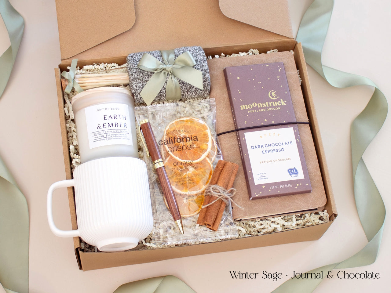 A thoughtful men’s gift box featuring a chocolate brown journal, pen, artisan chocolate bar, candle, white mug, and socks tied in sage ribbon, styled in a modern winter palette.