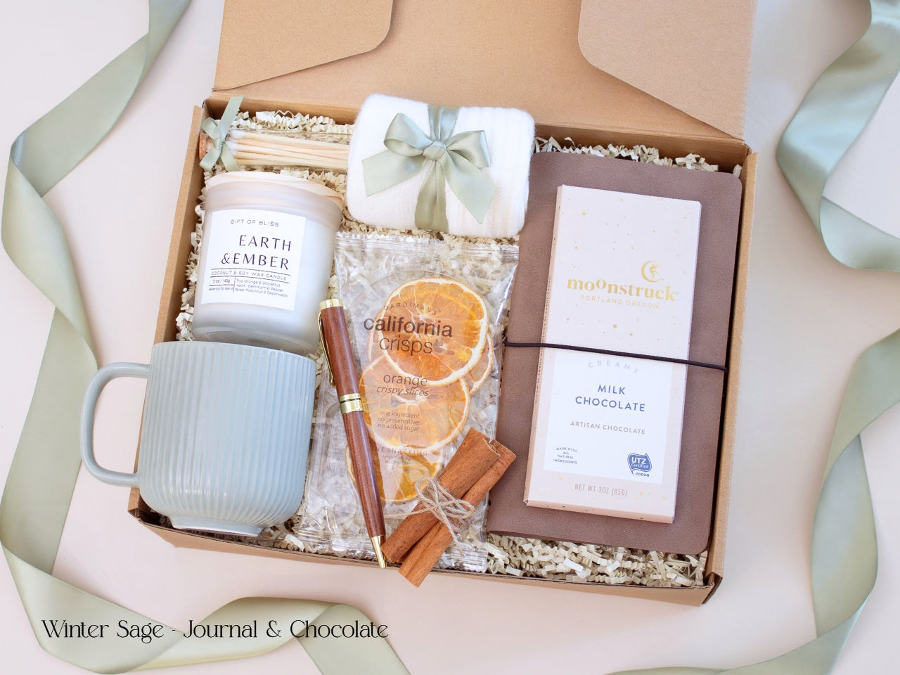 A thoughtful men’s gift box featuring a chocolate brown journal, pen, artisan chocolate bar, candle, white mug, and socks tied in sage ribbon, styled in a modern winter palette.