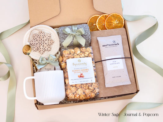 A cozy neutral-toned gift box featuring a caramel popcorn bag, artisan cocoa, mixed seasonal spices for mulling or simmering, brown journal, white mug, socks tied in sage ribbon, and decorative dried orange slices.