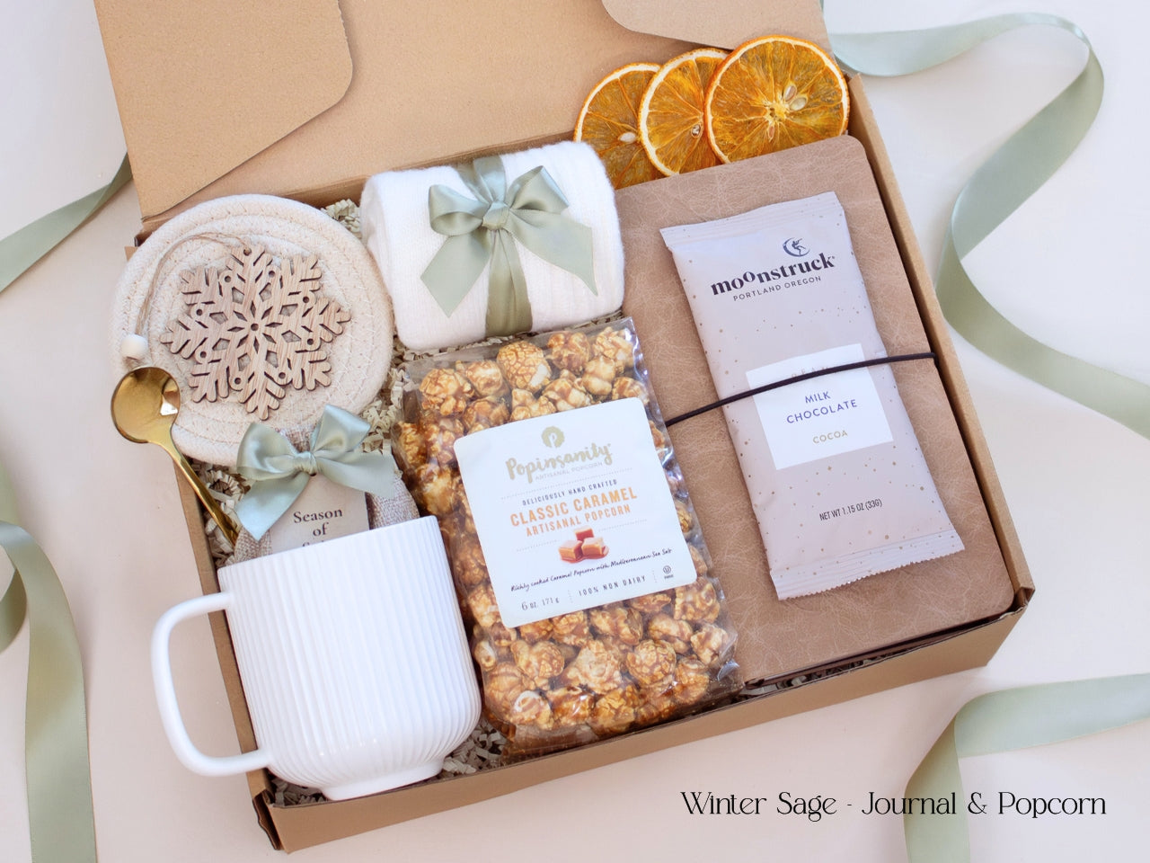 A cozy neutral-toned gift box featuring a caramel popcorn bag, artisan cocoa, mixed seasonal spices for mulling or simmering, brown journal, white mug, socks tied in sage ribbon, and decorative dried orange slices.