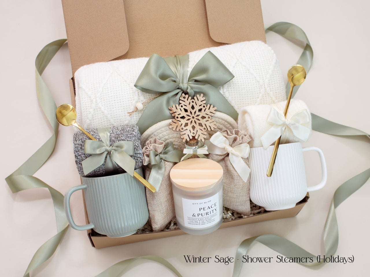 A serene holiday gift box for couples featuring a white knit blanket, two mugs, cozy socks, 2 bags of aromatherapy shower steamers, and a Peace & Purity candle, accented with a wooden snowflake ornament. The soothing sage green tones and ribbon evoke relaxation and festive calm.