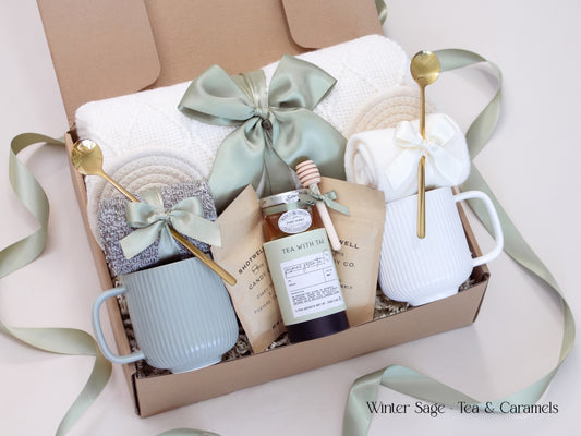 A cozy Winter Sage gift box for couples featuring a white knit blanket, two mugs, soft knit socks, Tea with Tae artisan tea, Shotwell caramels, and a wooden honey dipper. Tied with satin sage ribbons for an elegant, ready-to-gift presentation.
